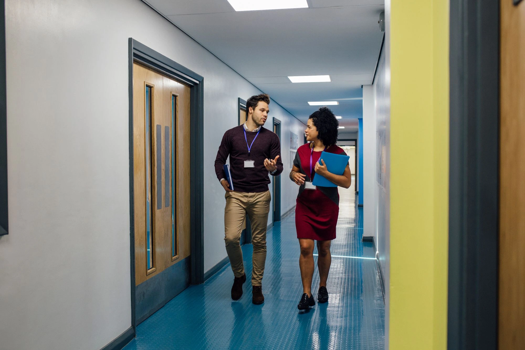 A male and female teacher walking down the school corridor wearing lanyards and having a discussion.