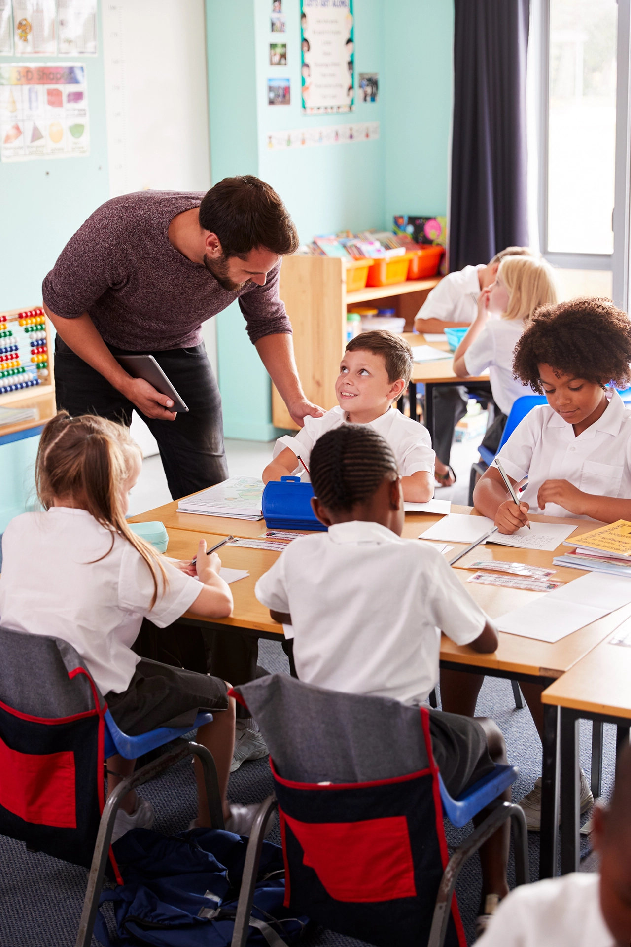 Male teacher leaning over desk of children during class.
