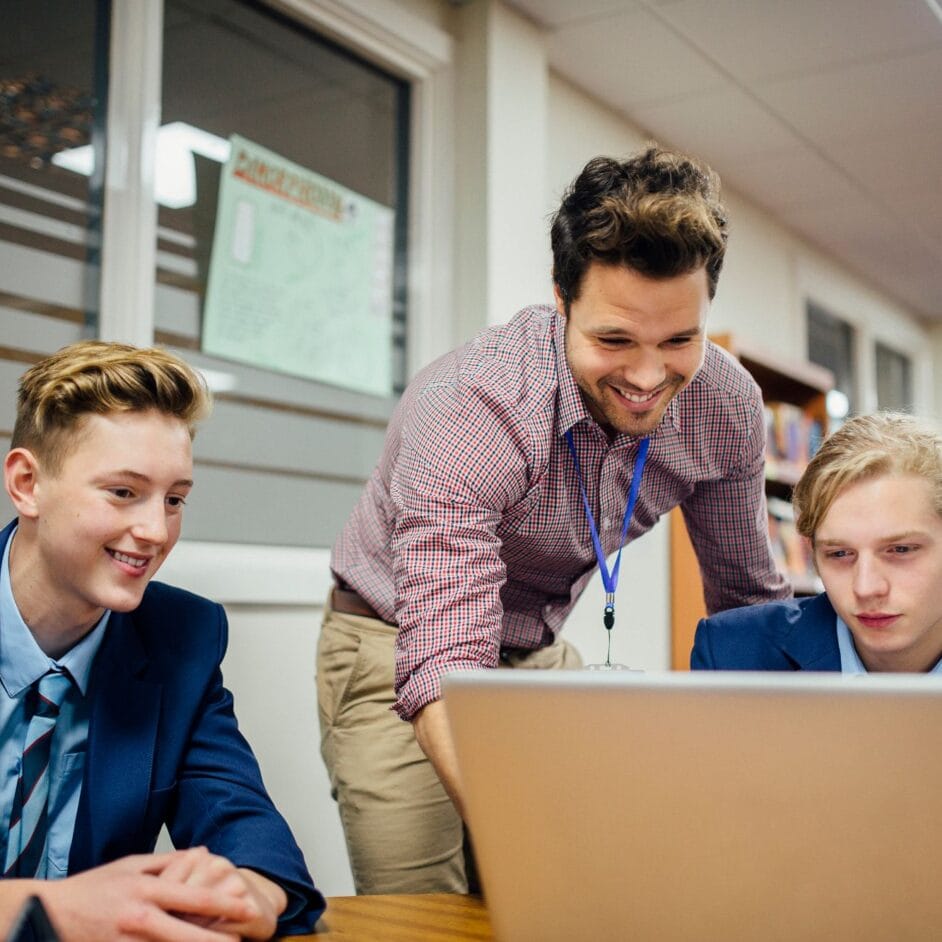 Teacher in pink shirt and blue lanyard leaning over desk to help students on laptops.