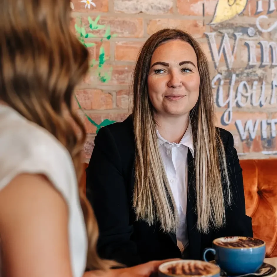 two females sitting at a table smiling and drinking coffee