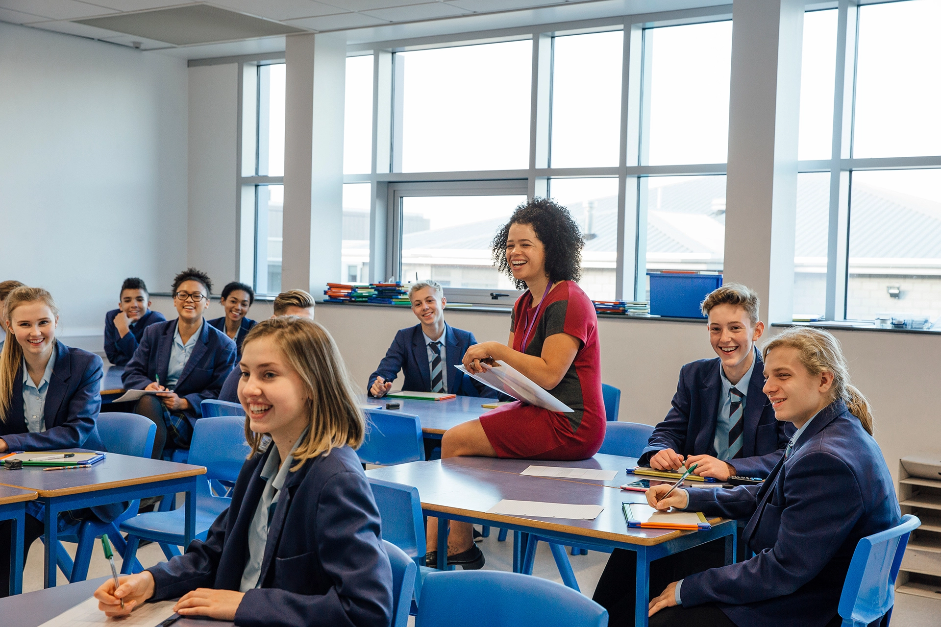 Teacher sits on desk and looks towards front of classroom during class discussion, with students at desks surrounding her.